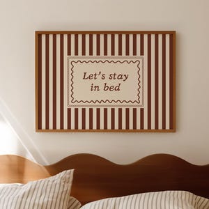 Puede incluir: Un cuadro rectangular enmarcado en madera con un fondo de rayas verticales marrones y blancas. En el centro, una etiqueta rectangular con borde ondulado contiene el texto marrón Let's stay in bed en una fuente serif sencilla.