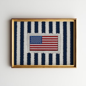 Puede incluir: Obra de arte enmarcada que presenta un diseño bordado de la bandera estadounidense. La bandera, con sus franjas rojas y blancas y su campo azul de estrellas, destaca. El fondo tiene un patrón de rayas azules y blancas, todo dentro de un marco dorado.