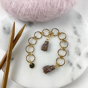 Könnte beinhalten: Ein goldenes Kettenarmband mit zwei lila Katzenanhängern. Das Armband liegt auf einer weißen Marmorfläche mit zwei Holzstricknadeln.