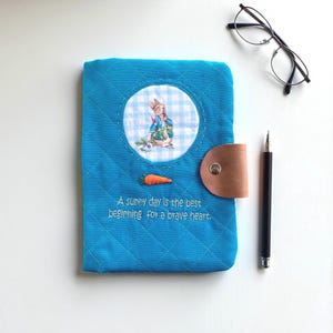 Può includere: Una copertina di diario trapuntata blu con un'illustrazione di Peter Rabbit e un ricamo di carota. La copertina ha una chiusura con cinturino in pelle marrone e il testo "A sunny day is the best beginning for a brave heart."