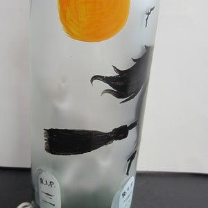 May include: A frosted glass bottle with a Halloween theme. The bottle features a hand-painted orange sun, a witch on a broom, and two tombstones with "R.I.P." on them. The base of the bottle is painted green.