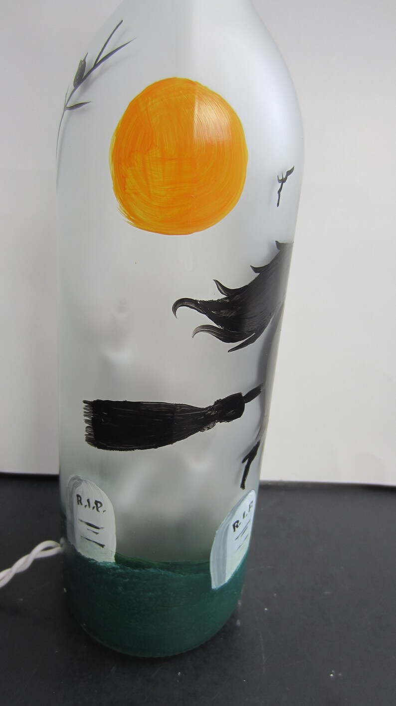 May include: A frosted glass bottle with a Halloween theme. The bottle features a hand-painted orange sun, a witch on a broom, and two tombstones with "R.I.P." on them. The base of the bottle is painted green.