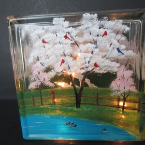 May include: A clear glass block lamp featuring a hand-painted spring scene. The artwork depicts a flowering tree with red and blue birds, a pond, and a wooden fence. The lamp is illuminated from within, creating a warm glow.