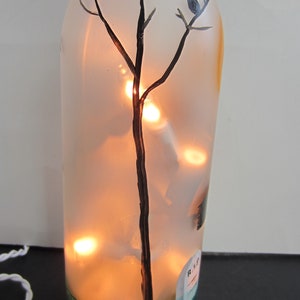 May include: A decorative glass bottle with a painted tree and a small bird, illuminated by warm white lights inside. The bottle has a green base and a small tombstone design. A white cord extends from the bottom.