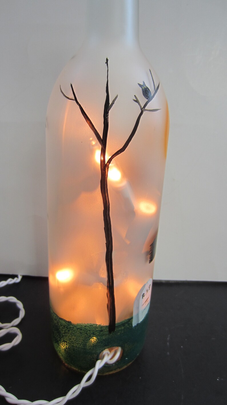 May include: A decorative glass bottle with a painted tree and a small bird, illuminated by warm white lights inside. The bottle has a green base and a small tombstone design. A white cord extends from the bottom.