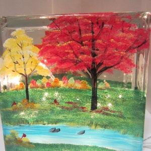 May include: A clear glass block with a hand-painted landscape scene. The artwork depicts a vibrant autumn scene with a red tree, a yellow tree, a blue stream, and green grass. Small lights are embedded within the block, illuminating the artwork.