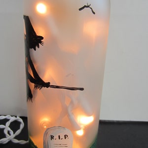 May include: A frosted glass bottle with interior lights, hand-painted with a Halloween theme. The design includes a witch on a broom, bats, and a tombstone with "R.I.P." on it. The base is painted green.