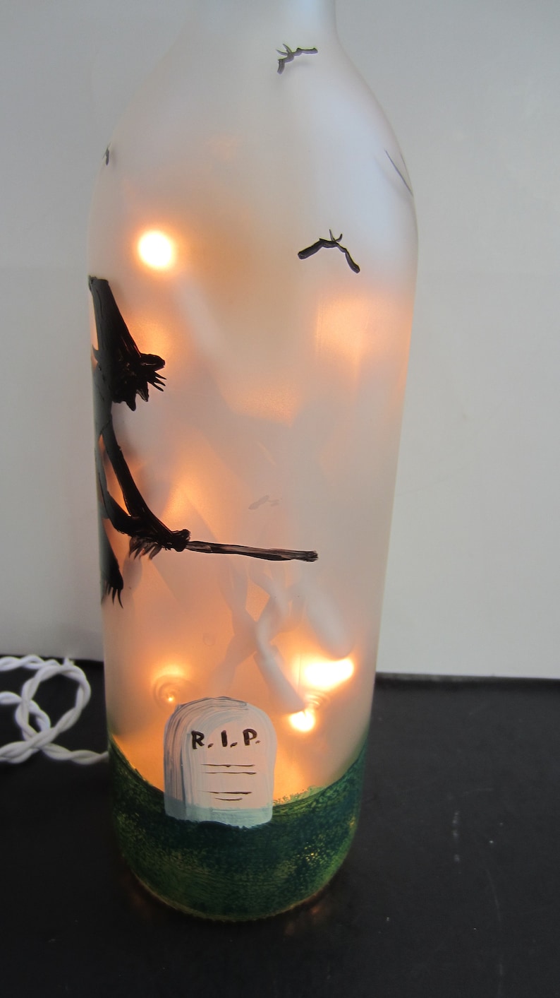 May include: A frosted glass bottle with interior lights, hand-painted with a Halloween theme. The design includes a witch on a broom, bats, and a tombstone with "R.I.P." on it. The base is painted green.