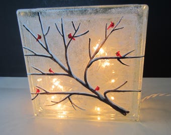 May include: A clear glass block with a winter scene painted on it. A dark brown tree branch with red cardinals is painted on the front. The block is lit from within by small, warm white lights.