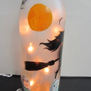 May include: A frosted glass bottle decorated with a Halloween theme. The bottle features a painted witch on a broomstick, a large orange moon, and two tombstones. The bottle is illuminated with warm white fairy lights, creating a festive glow.
