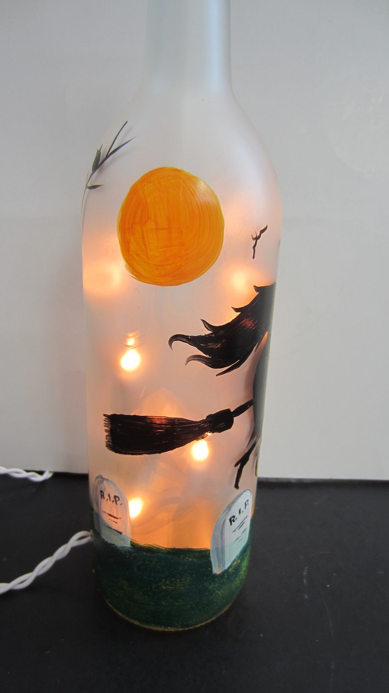 May include: A frosted glass bottle decorated with a Halloween theme. The bottle features a painted witch on a broomstick, a large orange moon, and two tombstones. The bottle is illuminated with warm white fairy lights, creating a festive glow.