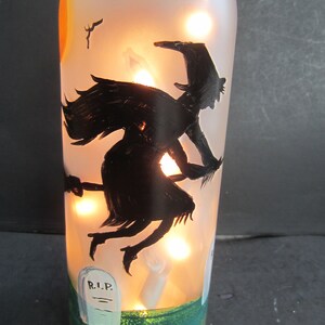May include: A frosted glass bottle with a Halloween theme. The bottle features a black silhouette of a witch flying on a broomstick, a graveyard scene, and a painted sunset. The bottle is illuminated with warm white lights.