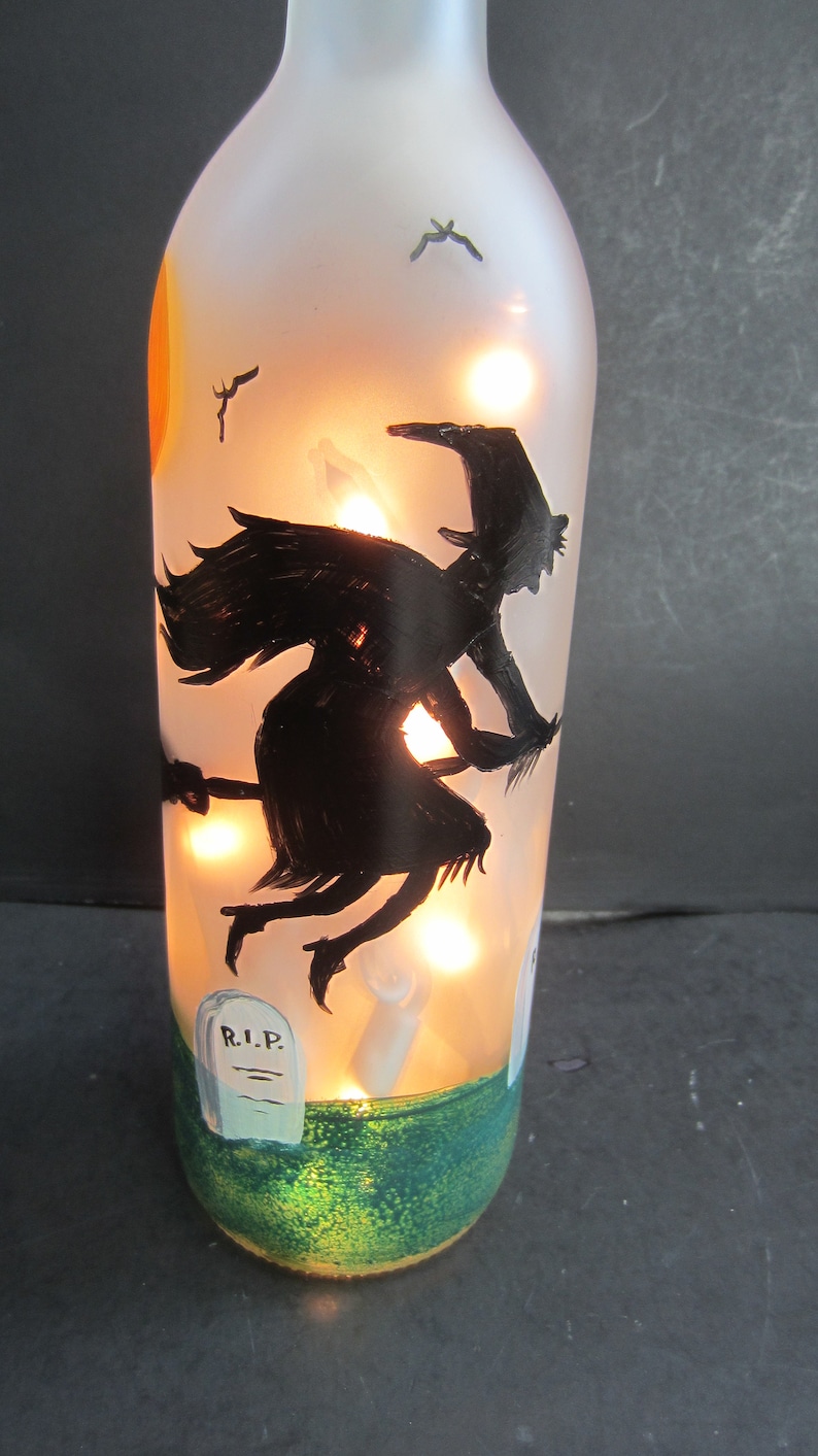 May include: A frosted glass bottle with a Halloween theme. The bottle features a black silhouette of a witch flying on a broomstick, a graveyard scene, and a painted sunset. The bottle is illuminated with warm white lights.