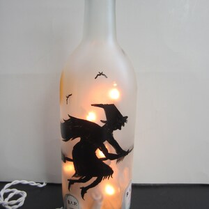 May include: A clear glass bottle with a frosted finish, illuminated by warm lights. A black silhouette of a witch on a broomstick is painted on the bottle, with a graveyard scene at the base. The bottle is decorated for Halloween.