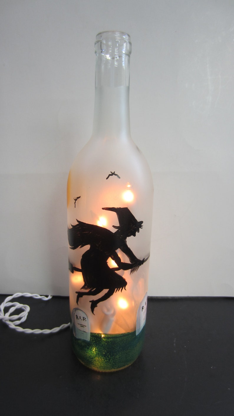 May include: A clear glass bottle with a frosted finish, illuminated by warm lights. A black silhouette of a witch on a broomstick is painted on the bottle, with a graveyard scene at the base. The bottle is decorated for Halloween.