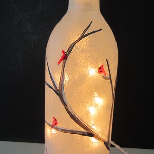 May include: A frosted glass bottle lamp with a winter scene. The bottle is decorated with painted branches and red cardinal birds. Small lights inside the bottle illuminate the design, creating a warm glow. A white cord extends from the base.
