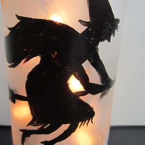 May include: A frosted glass bottle with a black silhouette of a witch flying on a broomstick. The bottle is illuminated from within, creating a warm glow. A tombstone with "R.I.P." is painted on the bottle, and a green base adds a decorative touch.