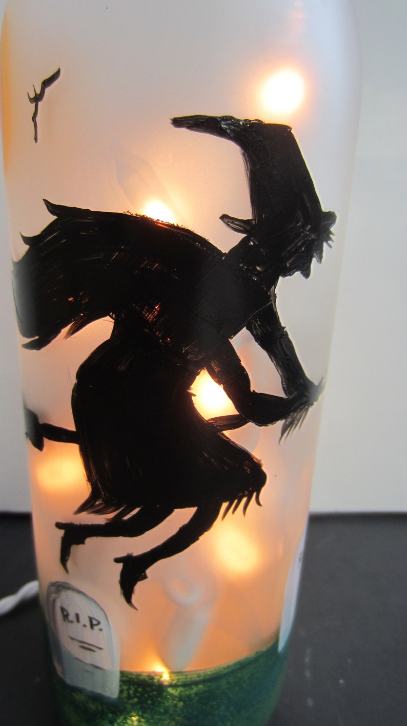 May include: A frosted glass bottle with a black silhouette of a witch flying on a broomstick. The bottle is illuminated from within, creating a warm glow. A tombstone with "R.I.P." is painted on the bottle, and a green base adds a decorative touch.