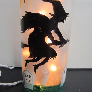 May include: A Halloween-themed decorative bottle with a black silhouette of a witch flying on a broomstick. The bottle is illuminated with warm lights, and features painted tombstones and a green base. Small black bats are also painted on the bottle.