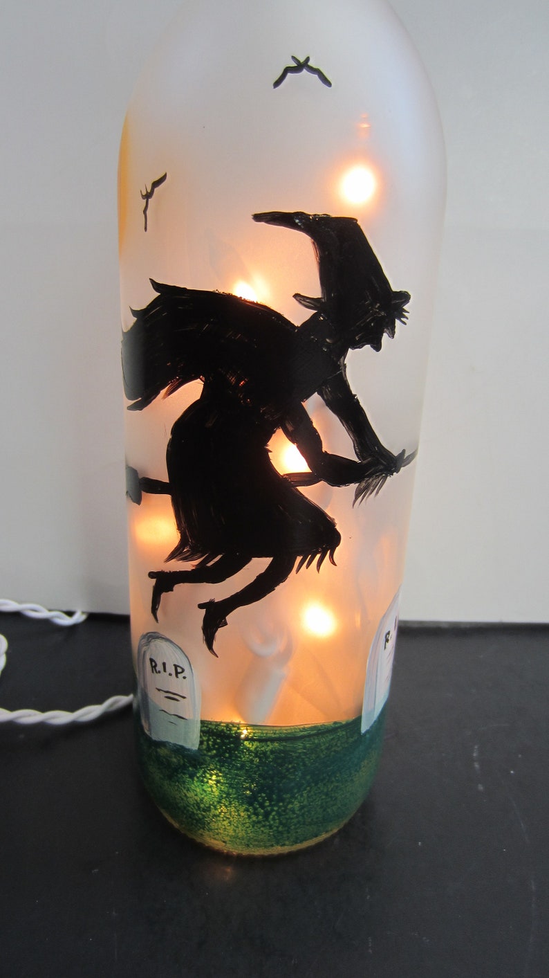 May include: A Halloween-themed decorative bottle with a black silhouette of a witch flying on a broomstick. The bottle is illuminated with warm lights, and features painted tombstones and a green base. Small black bats are also painted on the bottle.