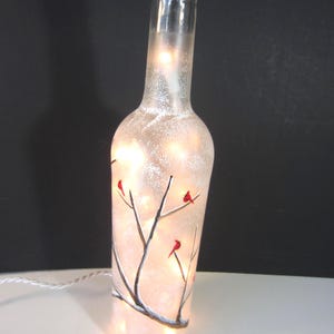 May include: A clear glass bottle with a frosted finish, illuminated by small white lights inside. The bottle is decorated with a painted winter scene of bare tree branches and red birds. A white cord extends from the base.