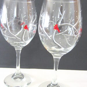 May include: Two clear glass wine glasses featuring hand-painted winter scenes. Each glass displays white snow-covered tree branches and small red cardinal birds with yellow beaks, accented by white dots representing falling snow against the glass.