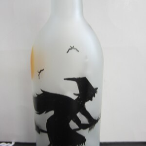 May include: Frosted glass bottle with a black silhouette of a witch on a broomstick, flying in front of a yellow sun. The bottle has a green base and a white cord.