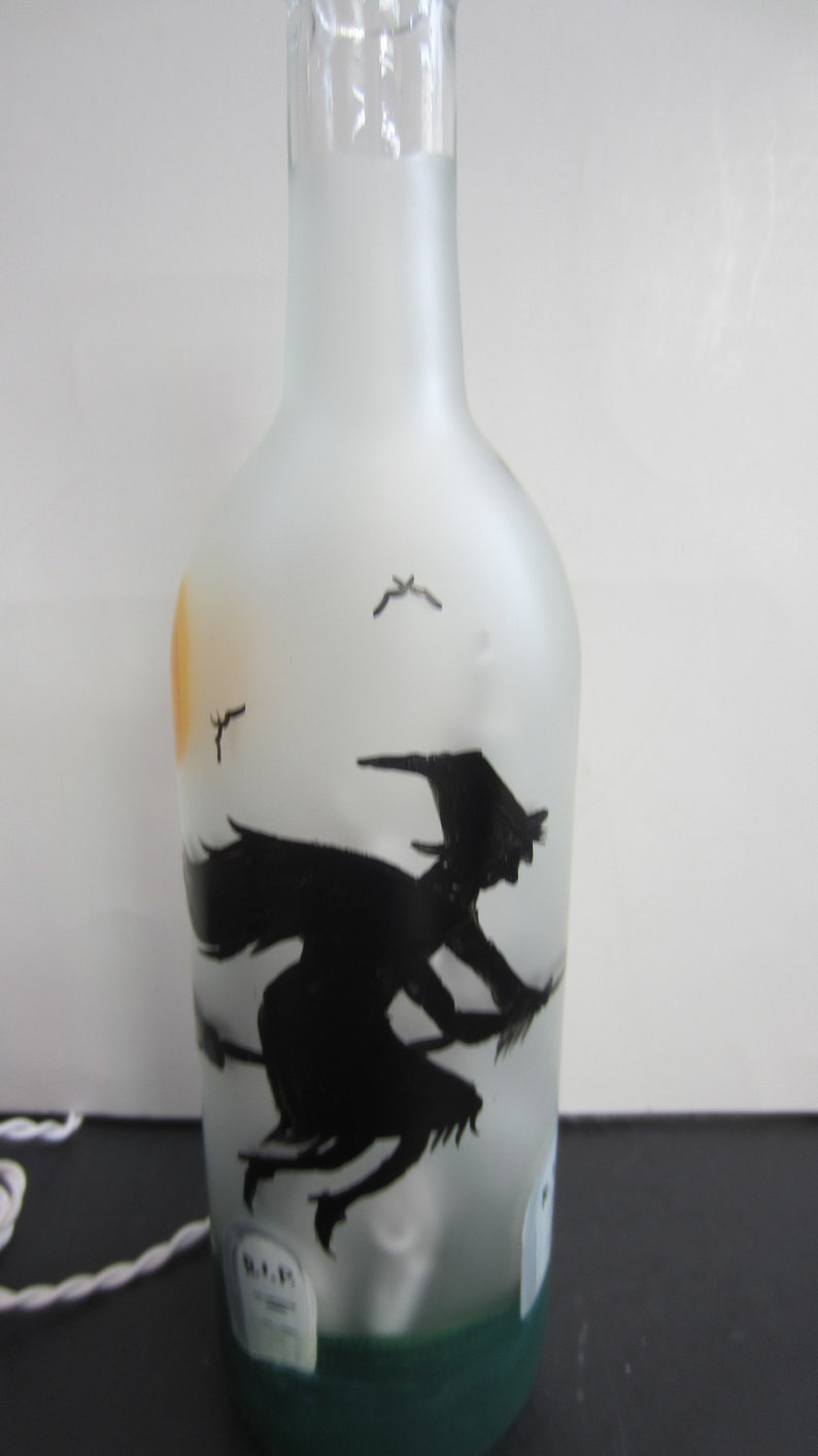 May include: Frosted glass bottle with a black silhouette of a witch on a broomstick, flying in front of a yellow sun. The bottle has a green base and a white cord.