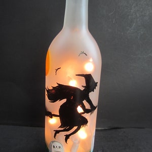 May include: A frosted glass bottle with a Halloween theme. The bottle features a black silhouette of a witch flying on a broom, a full moon, and a graveyard scene. The bottle is illuminated with small lights.