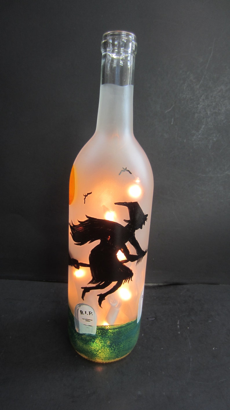 May include: A frosted glass bottle with a Halloween theme. The bottle features a black silhouette of a witch flying on a broom, a full moon, and a graveyard scene. The bottle is illuminated with small lights.