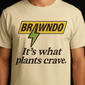 Puede incluir: Camiseta color crema con la palabra "BRAWNDO" en un rectángulo amarillo y negro, un rayo verde y el texto "It's what plants crave." Una camiseta informal de manga corta.
