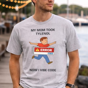 Puede incluir: Camiseta gris claro con el texto "MY MOM TOOK TYLENOL" sobre un dibujo animado de una persona corriendo con un letrero de "ERROR". El texto "NOW I VIBE CODE" está impreso debajo.