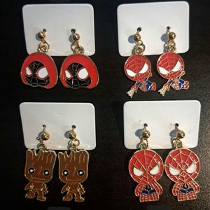 May include: Four pairs of gold-tone earrings with different cartoon character designs. The earrings are displayed on white cards with numbers 1 through 4. The designs include a red and black Spider-Man, a brown Groot, and a red Spider-Man with a blue and white suit.