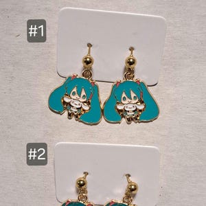 May include: Gold-coloured dangle earrings with a teal-haired anime character design. The earrings have a gold ball at the top and a detailed image of the character with teal hair and a white outfit. The earrings are displayed on white cards.