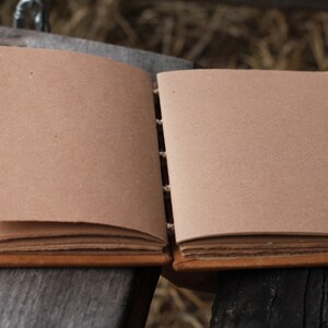 Puede incluir: Diario artesanal abierto con p&aacute;ginas en blanco de color marr&oacute;n claro. El diario tiene una cubierta y lomo de cuero marr&oacute;n, con costuras visibles. Las p&aacute;ginas son gruesas y parecen estar hechas de papel reciclado. El diario est&aacute; abierto sobre una superficie de madera envejecida.