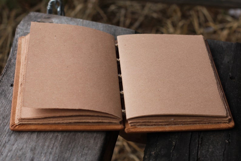Puede incluir: Diario artesanal abierto con p&aacute;ginas en blanco de color marr&oacute;n claro. El diario tiene una cubierta y lomo de cuero marr&oacute;n, con costuras visibles. Las p&aacute;ginas son gruesas y parecen estar hechas de papel reciclado. El diario est&aacute; abierto sobre una superficie de madera envejecida.