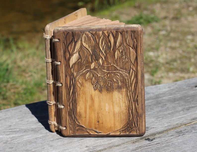 Puede incluir: Un diario de madera hecho a mano con un dise&ntilde;o de &aacute;rbol tallado en la portada. El diario est&aacute; encuadernado con hilo y tiene una est&eacute;tica r&uacute;stica y natural. Las p&aacute;ginas son visibles y la madera tiene un tono marr&oacute;n c&aacute;lido.
