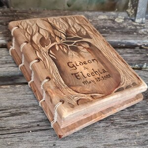 Puede incluir: Un libro de madera con un dise&ntilde;o de &aacute;rbol tallado en la portada. El libro est&aacute; encuadernado con hilo blanco y presenta los nombres "Gideon & Fletchia" y la fecha "May 25, 2022" grabados en la parte delantera. El libro es de madera marr&oacute;n claro.