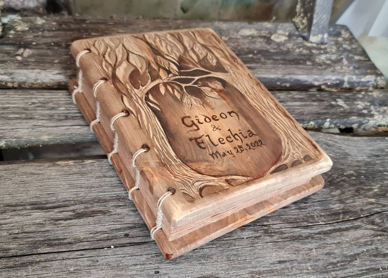 Puede incluir: Un libro de madera con un dise&ntilde;o de &aacute;rbol tallado en la portada. El libro est&aacute; encuadernado con hilo blanco y presenta los nombres "Gideon & Fletchia" y la fecha "May 25, 2022" grabados en la parte delantera. El libro es de madera marr&oacute;n claro.
