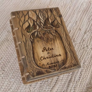 Puede incluir: Un diario de madera con un dise&ntilde;o de &aacute;rbol tallado en la portada. La portada est&aacute; grabada con los nombres "Peter & Christina" y la fecha "13 de noviembre de 2021". El diario est&aacute; encuadernado con costuras visibles.