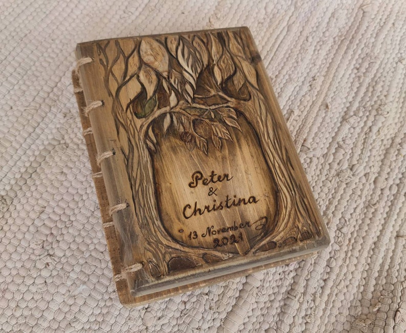 Puede incluir: Un diario de madera con un dise&ntilde;o de &aacute;rbol tallado en la portada. La portada est&aacute; grabada con los nombres "Peter & Christina" y la fecha "13 de noviembre de 2021". El diario est&aacute; encuadernado con costuras visibles.