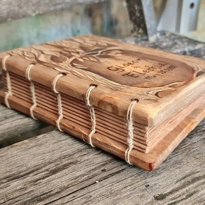 Puede incluir: Un libro de madera hecho a mano con un grabado de un &aacute;rbol en la portada. La portada tambi&eacute;n presenta los nombres "Gideon & Elechia" y la fecha "May 25 2023". Las p&aacute;ginas est&aacute;n encuadernadas con hilo. La madera tiene un tono c&aacute;lido y natural.