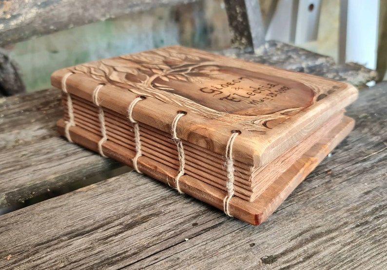 Puede incluir: Un libro de madera hecho a mano con un grabado de un &aacute;rbol en la portada. La portada tambi&eacute;n presenta los nombres "Gideon & Elechia" y la fecha "May 25 2023". Las p&aacute;ginas est&aacute;n encuadernadas con hilo. La madera tiene un tono c&aacute;lido y natural.