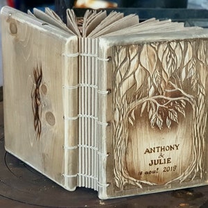 Puede incluir: Diario de madera hecho a mano con un dise&ntilde;o de &aacute;rbol tallado en la portada. El diario est&aacute; encuadernado con hilo blanco y presenta los nombres "ANTHONY & JULIE" y la fecha "16 aout 2019" tallados en la madera. El diario est&aacute; abierto.