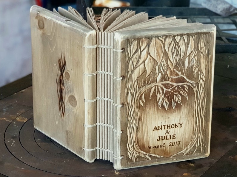 Puede incluir: Diario de madera hecho a mano con un dise&ntilde;o de &aacute;rbol tallado en la portada. El diario est&aacute; encuadernado con hilo blanco y presenta los nombres "ANTHONY & JULIE" y la fecha "16 aout 2019" tallados en la madera. El diario est&aacute; abierto.