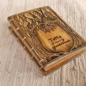 Puede incluir: Un libro de visitas de madera con un dise&ntilde;o de &aacute;rbol tallado en la portada. La portada es de color marr&oacute;n claro con detalles en marr&oacute;n oscuro y el texto "Totte Family Guestbook". Las p&aacute;ginas son visibles y de color beige claro. El libro est&aacute; encuadernado con hilo.