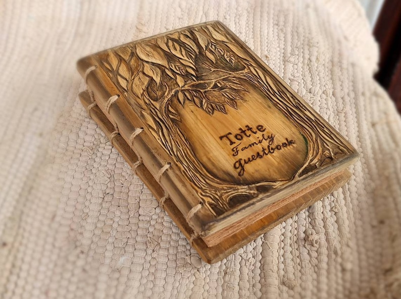 Puede incluir: Un libro de visitas de madera con un dise&ntilde;o de &aacute;rbol tallado en la portada. La portada es de color marr&oacute;n claro con detalles en marr&oacute;n oscuro y el texto "Totte Family Guestbook". Las p&aacute;ginas son visibles y de color beige claro. El libro est&aacute; encuadernado con hilo.