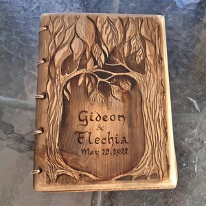 Puede incluir: Un diario de madera con un dise&ntilde;o de &aacute;rbol tallado en la portada. El diario est&aacute; encuadernado con anillas met&aacute;licas y presenta los nombres "Gideon & Elechia" y la fecha "May 25, 2022" tallados en la madera.