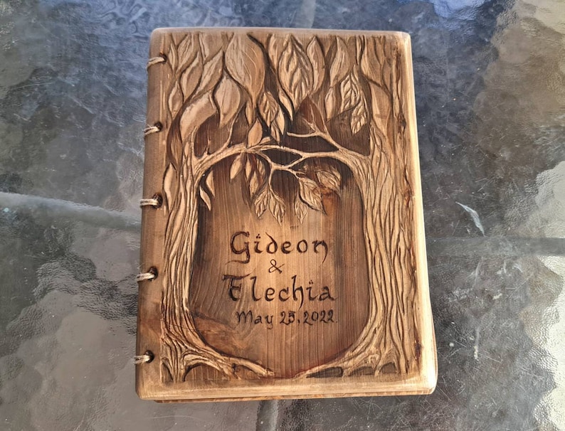 Puede incluir: Un diario de madera con un dise&ntilde;o de &aacute;rbol tallado en la portada. El diario est&aacute; encuadernado con anillas met&aacute;licas y presenta los nombres "Gideon & Elechia" y la fecha "May 25, 2022" tallados en la madera.