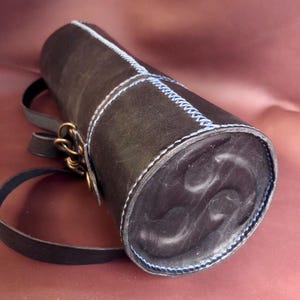 May include: Black leather cylindrical bag with a blue stitched design and a long strap. The bag has a metal clasp.